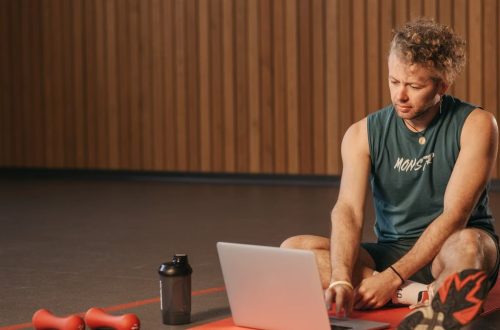 Man sitting on exercise mat with laptop, surrounded by dumbbells, creates a unique fitness workspace.
